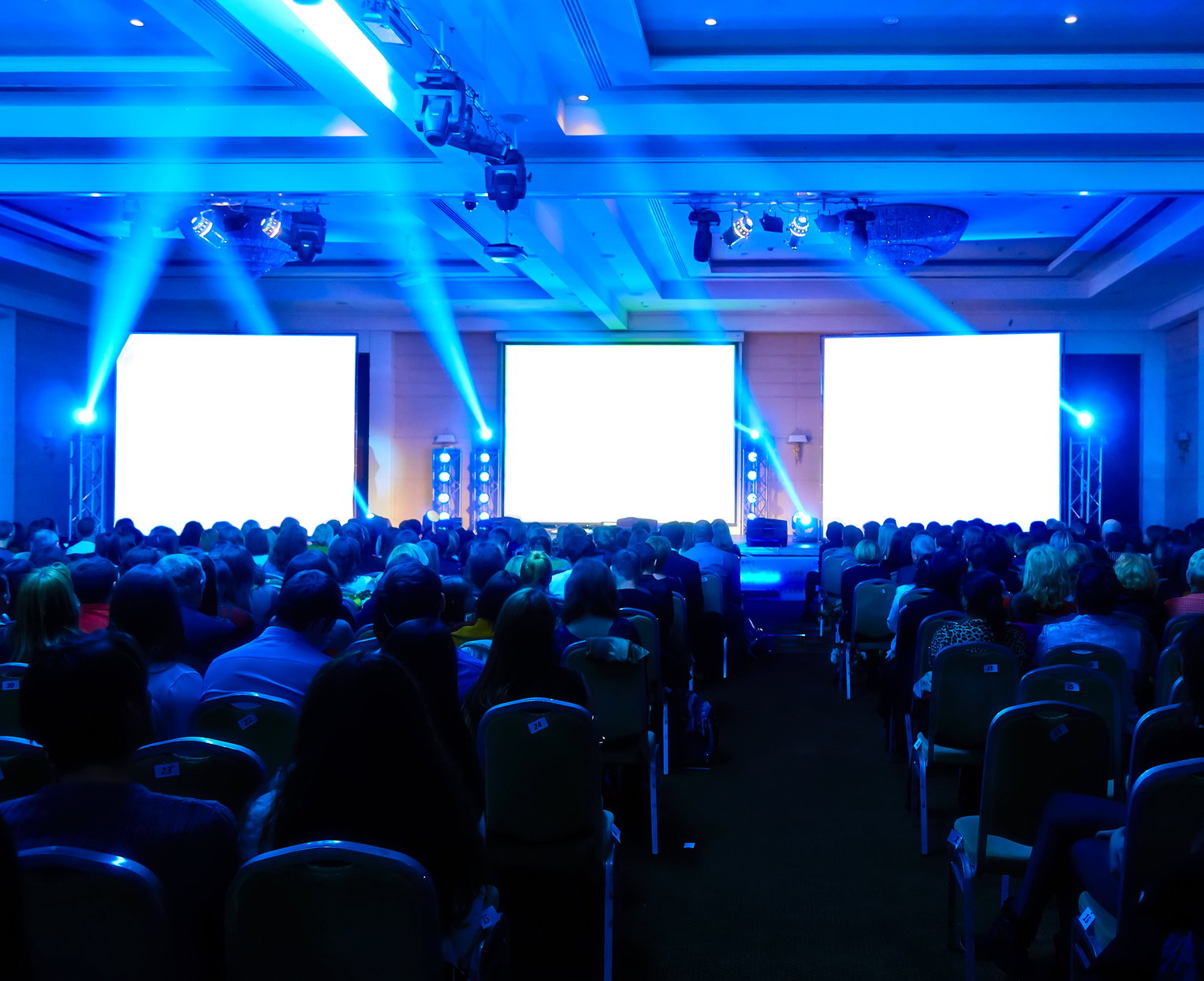 A view of people sitting at an event facing the three LCD panels on the stage